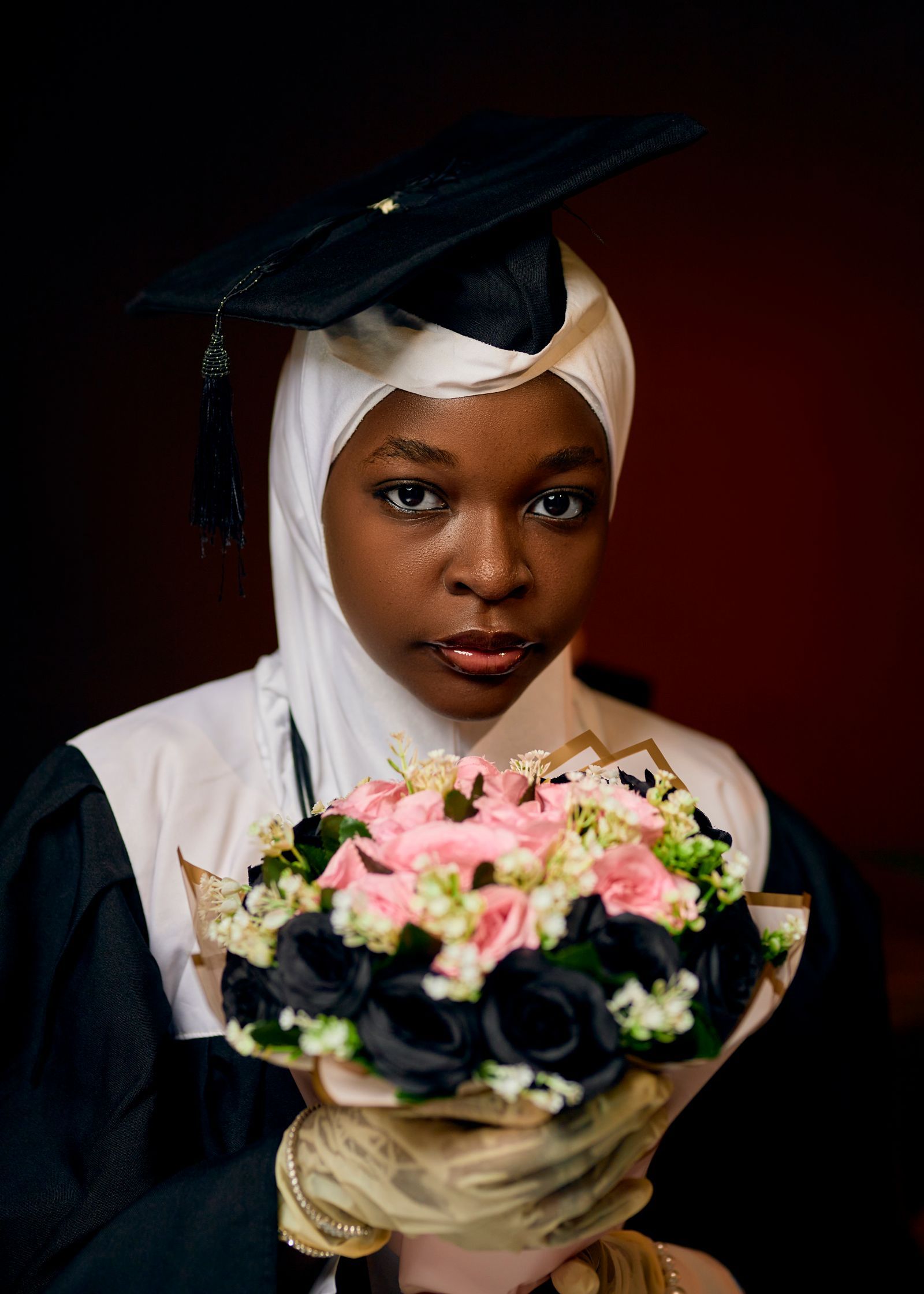 Graduate in a cap and hijab holding flowers in a refined studio portrait.