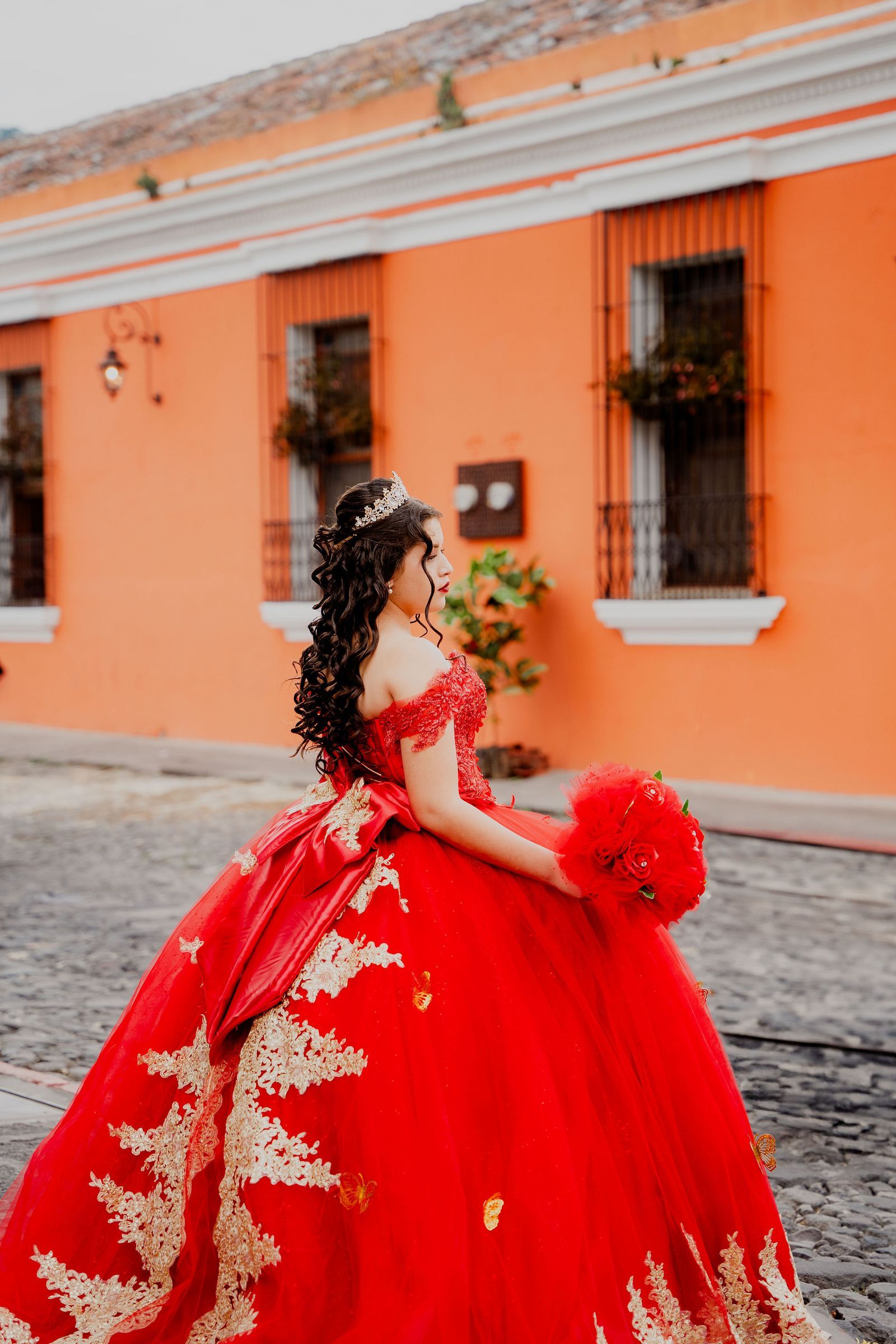 Quinceañera in a red gown holding a bouquet on a colorful historic street.