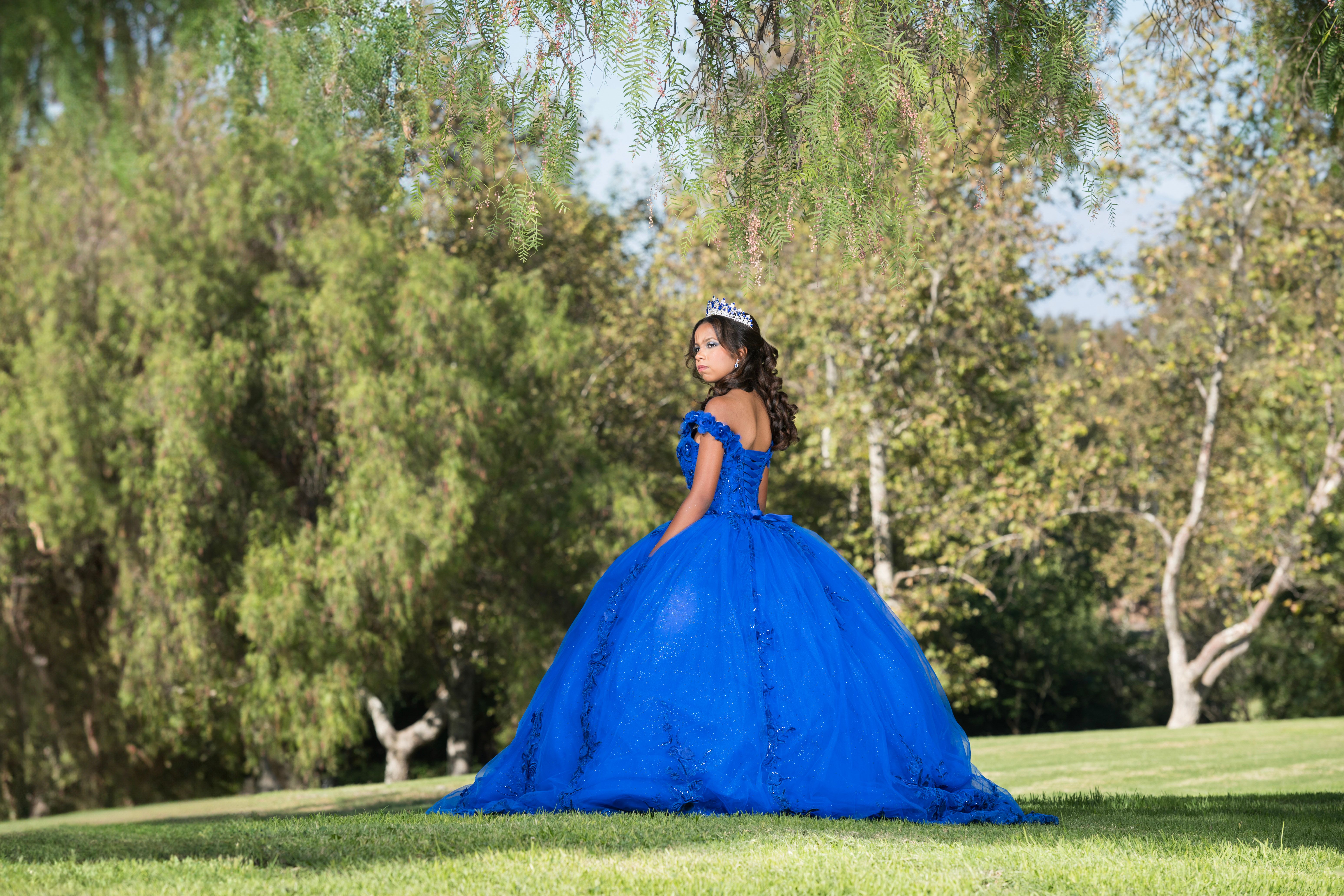 Quinceañera in a royal-blue gown posing on a sunlit lawn.