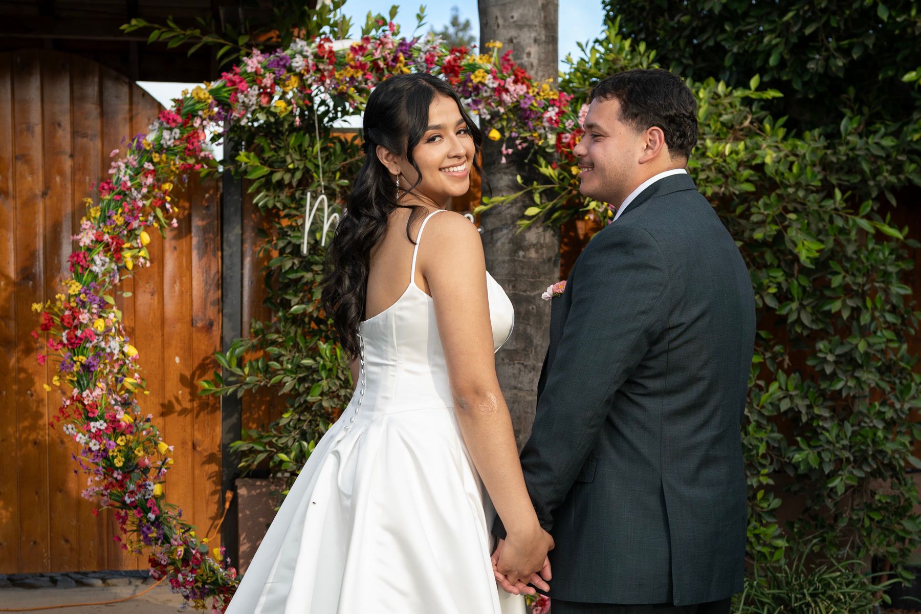 Newlyweds smiling at one another beside a colorful floral doorway.