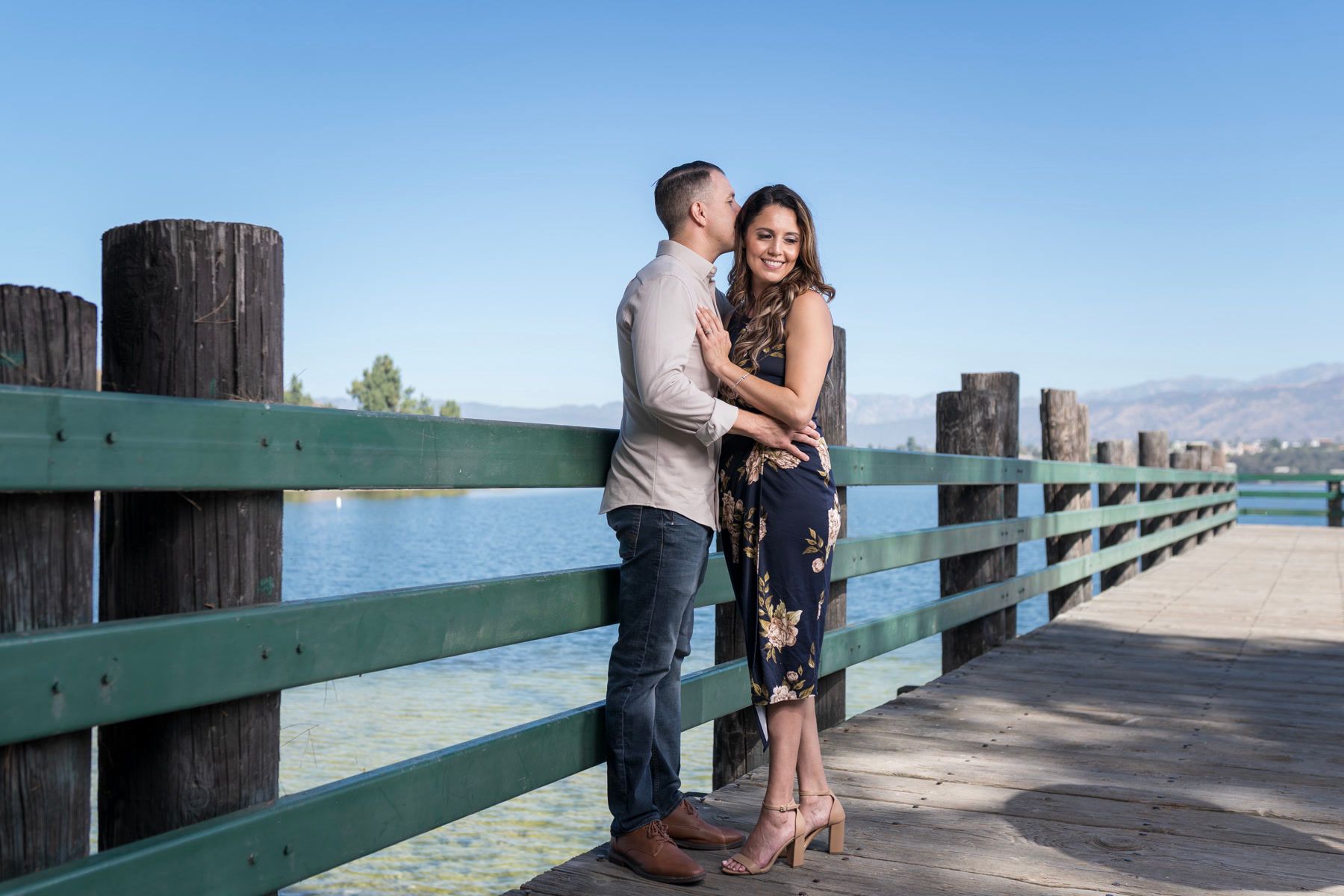 Engaged couple embracing beside the railing of a lakeside wooden pier.