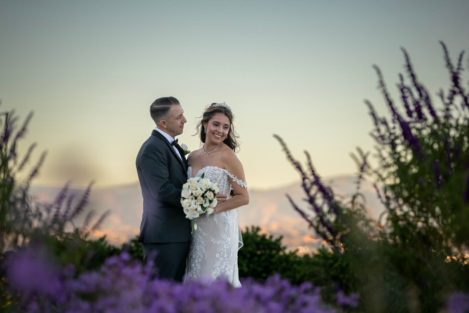 Newlyweds standing together behind purple flowers at sunset.